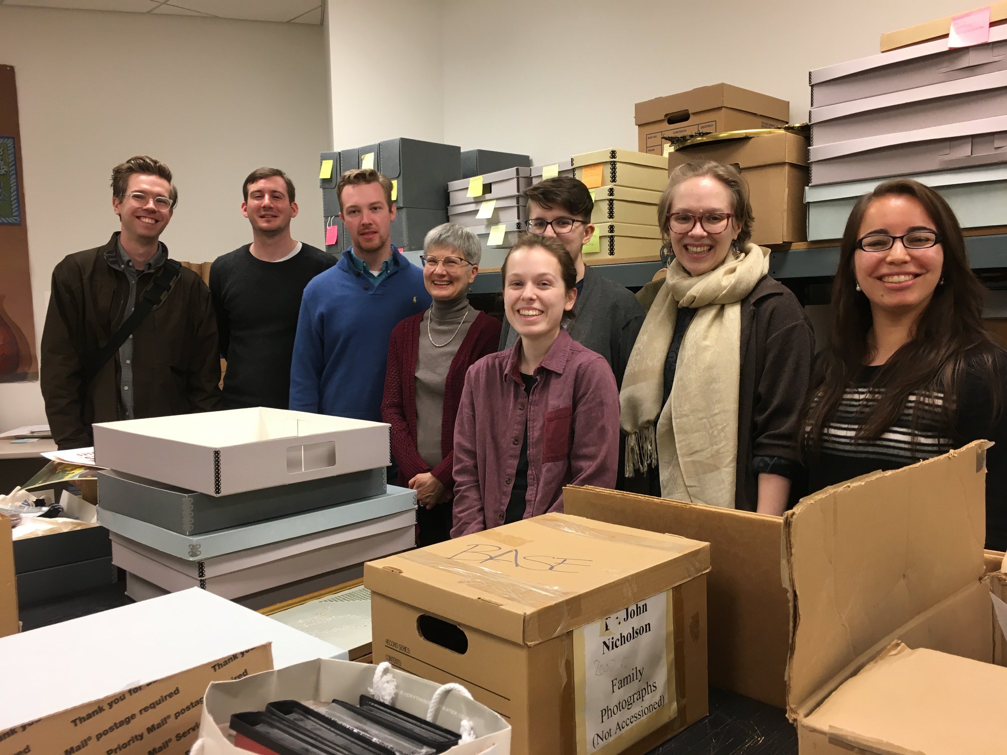 Seven students and an archivist stand together in a room full of archival boxes,